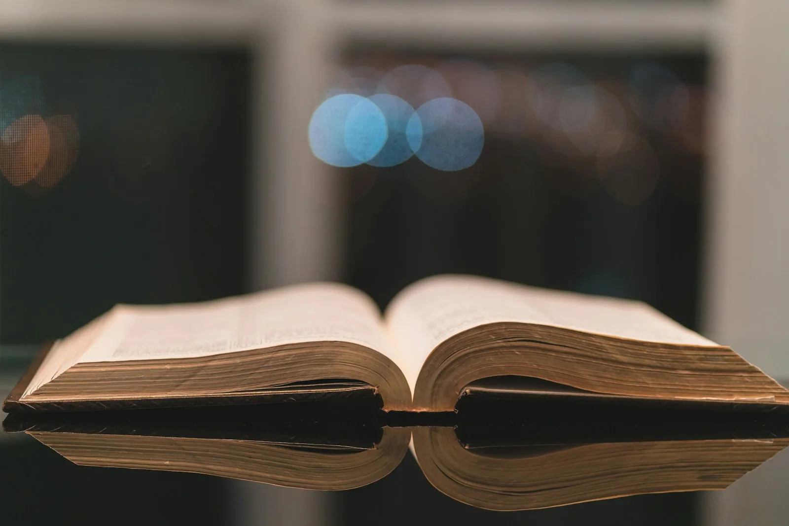 Livro antigo aberto sobre uma mesa escura, com uma biblioteca desfocada ao fundo